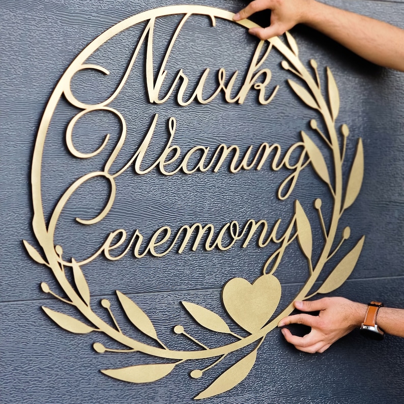 May include: A gold-colored wooden sign with a wreath design and the words "Nuwk Wedding Ceremony" in a cursive font. A heart shape is in the center of the wreath.