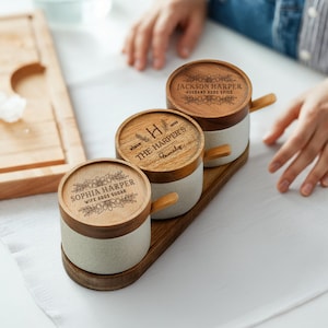 May include: Three ceramic spice jars with wooden lids and handles, on a wooden tray. The lids are engraved with floral designs and personalized text: "Sophia Harper Wife Adds Sugar", "The Harper's Family", and "Jackson Harper Husband Adds Spice".