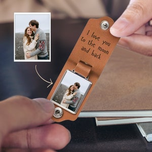Könnte beinhalten: Ein brauner Leder-Schlüsselanhänger mit einem silbernen Fotorahmen. Der Fotorahmen enthält ein Bild eines Paares. Der Schlüsselanhänger trägt die Gravur "I love you to the moon and back".