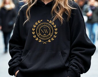May include: A black hooded sweatshirt featuring a gold-colored circular emblem on the chest. The emblem displays a laurel wreath surrounding the letters NW, the text NICOLE WILLIAMS, and the text ESTD 1985 with three small stars above the letters.