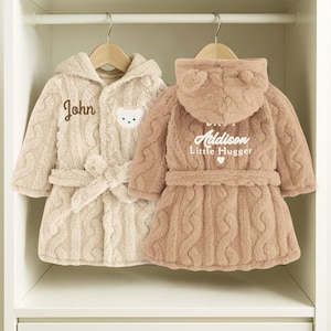 May include: Two plush, hooded children's bathrobes in a closet. One is beige with "John" embroidered on it and a bear face applique. The other is a light brown color with "Addison Little Hugger" printed on the back. Both have a cable knit design and a matching belt.
