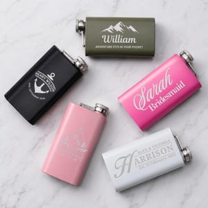 May include: Five rectangular metal flasks in olive green, black, hot pink, light pink, and white. Each flask features a silver screw-top cap and custom laser-engraved text and graphics, including names, dates, and designs like an anchor and mountains.