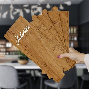 Könnte beinhalten: Ein hölzerner Wachstumsmesser mit Puzzle-Design. Die Karte ist mit dem Namen "Juliette Wilson" personalisiert und zeigt ein Blumenmuster. Die Karte misst bis zu 152,4 cm.