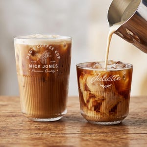 Puede incluir: Dos vasos de cristal estriado llenos de café con hielo. El vaso más alto tiene el texto blanco DAD'S COFFEE CUP, ESTD 2024, NICK JONES, Premium Quality, y un gráfico de planta de café. El vaso más bajo tiene el nombre Juliette en letra cursiva blanca.