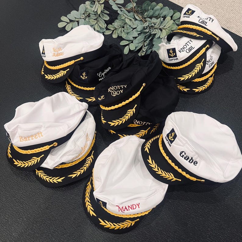 May include: An assortment of white and black captain hats with gold embroidered details. Some hats have names like "Barrett", "Mandy", and "Gabe" embroidered in gold. Other hats have the words "Captain", "Knotty Boy", and "Knotty Girl".