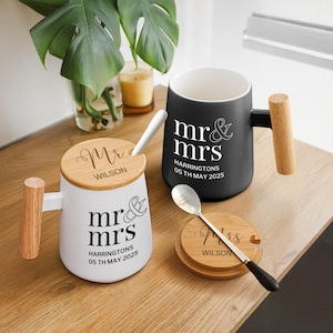Op de afbeelding: Witte en zwarte keramische mokken met houten handvatten en deksels. De mokken zijn gepersonaliseerd met "mr & mrs", een datum en een naam. Een lepel en een onderzetter zijn ook inbegrepen. De mokken staan op een houten oppervlak.