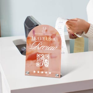 Op de afbeelding: Een roze acryl bord met een afgeronde bovenkant en een witte rand. Het bord zegt "Leave us a review" en heeft een QR-code en vijf sterren. Het bord staat op een wit aanrecht.