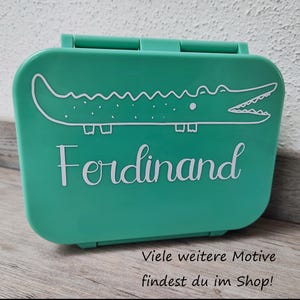 Könnte beinhalten: Eine grüne Lunchbox mit einem weißen Alligator-Motiv und dem Namen "Ferdinand" auf der Vorderseite.