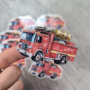 Könnte beinhalten: Ein roter Feuerwehrauto-Aufkleber mit einer gelben Leiter und einem Schlauch. Der Aufkleber ist auf einem transparenten Hintergrund.