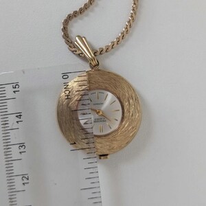 May include: A gold-toned pendant watch with a textured, circular face and a gold chain. The watch face is silver with gold hands and markers. The watch measures approximately 1.25 inches in diameter.