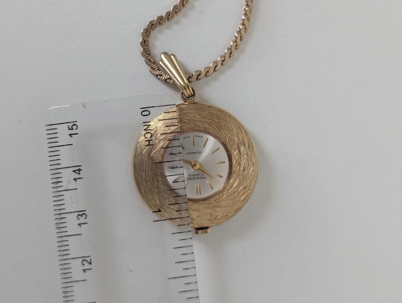 May include: A gold-toned pendant watch with a textured, circular face and a gold chain. The watch face is silver with gold hands and markers. The watch measures approximately 1.25 inches in diameter.