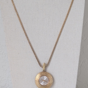 May include: A gold-toned pendant watch necklace. The watch face is white with gold-toned hands and markers. The pendant has a textured, circular design. The chain is a twisted rope style. The watch is displayed on a white surface.