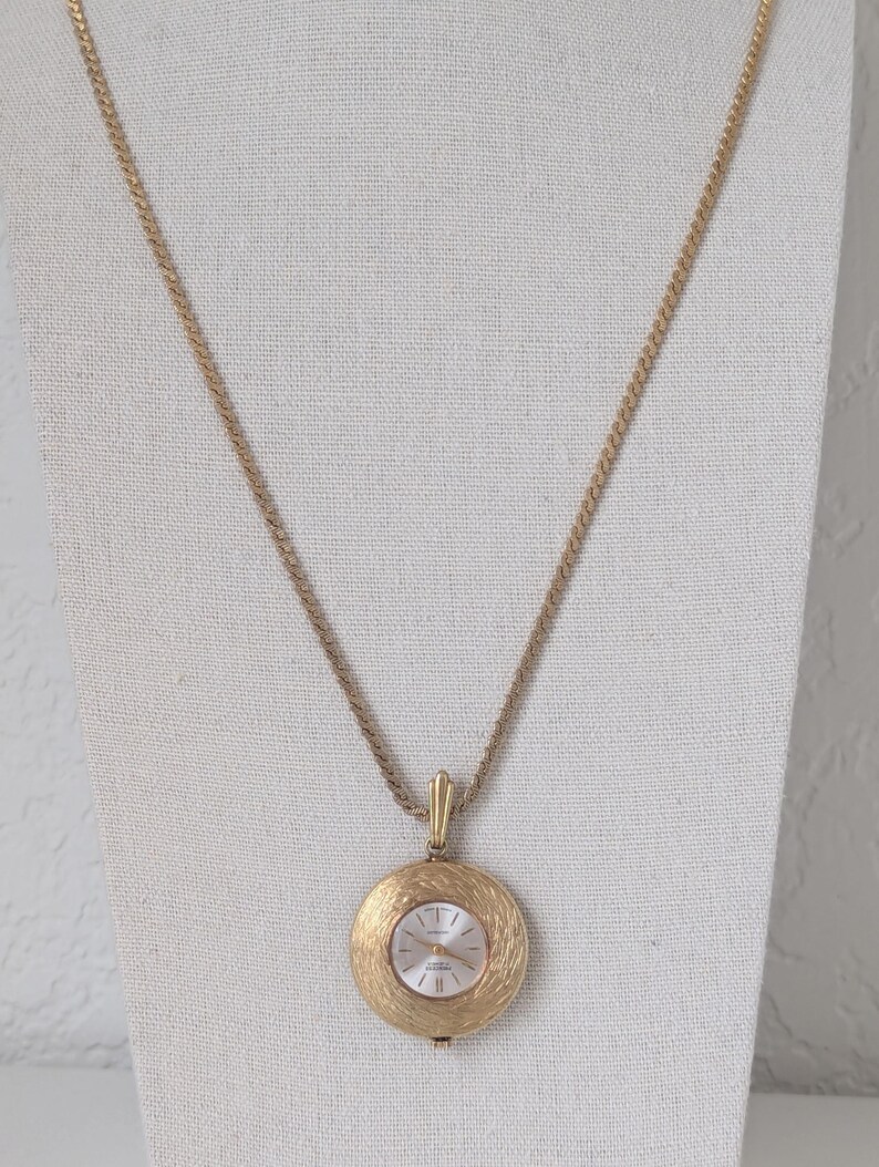 May include: A gold-toned pendant watch necklace. The watch face is white with gold-toned hands and markers. The pendant has a textured, circular design. The chain is a twisted rope style. The watch is displayed on a white surface.