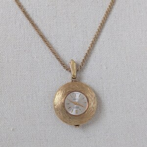 May include: A gold-toned pendant watch necklace. The round watch face has a textured gold frame and a white face with gold hands and markers. The chain is a delicate, twisted design.