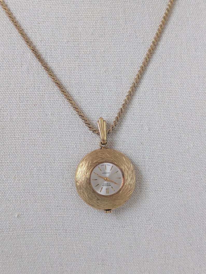 May include: A gold-toned pendant watch necklace. The round watch face has a textured gold frame and a white face with gold hands and markers. The chain is a delicate, twisted design.