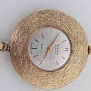 May include: A gold-toned Princess 17 Jewels Swiss Made pendant watch. The watch face is silver with gold hands and markers. The watch has a textured, circular frame and is attached to a chain.