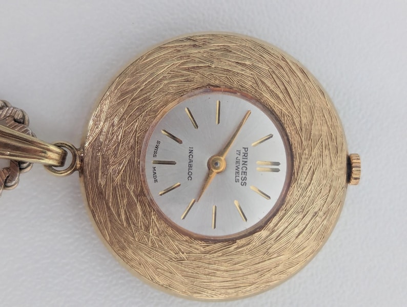 May include: A gold-toned Princess 17 Jewels Swiss Made pendant watch. The watch face is silver with gold hands and markers. The watch has a textured, circular frame and is attached to a chain.