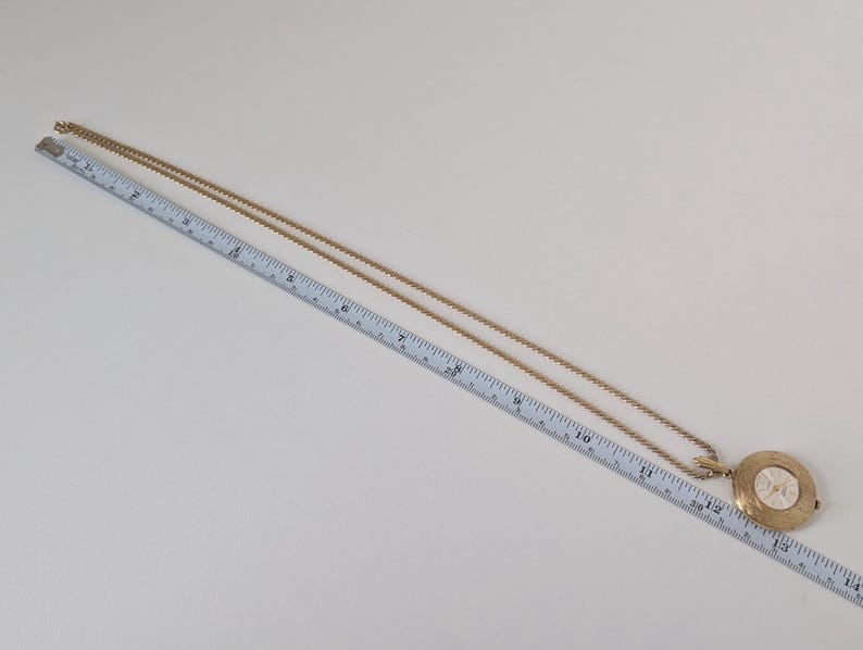 May include: Gold-toned pendant necklace with a round, ornate pendant. The pendant features a white center with a design. The chain is long and delicate. The necklace is approximately 14 inches long.