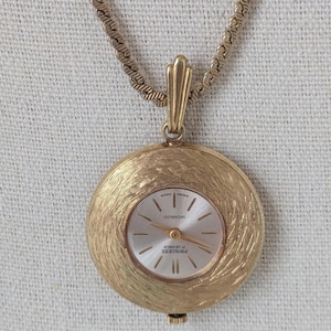 May include: A gold-toned pendant watch with a textured, circular face and a matching chain. The watch face is silver with gold hands and markers. The watch is marked "Princess 17 Jewels" and "Swiss Made".