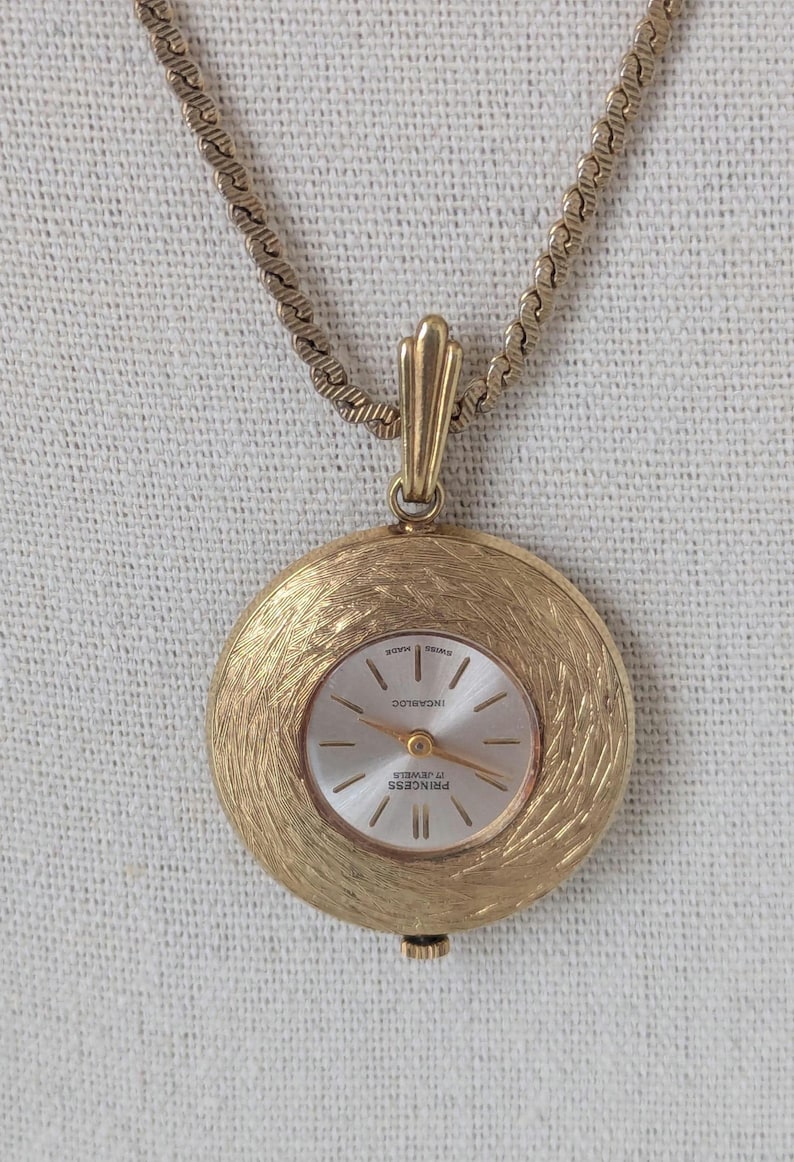 May include: A gold-toned pendant watch with a textured, circular face and a matching chain. The watch face is silver with gold hands and markers. The watch is marked "Princess 17 Jewels" and "Swiss Made".