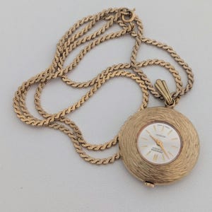 May include: A gold-toned pendant watch with a textured, circular face and a matching chain. The watch face has a white background with gold hands and markers. The chain is a twisted rope design.