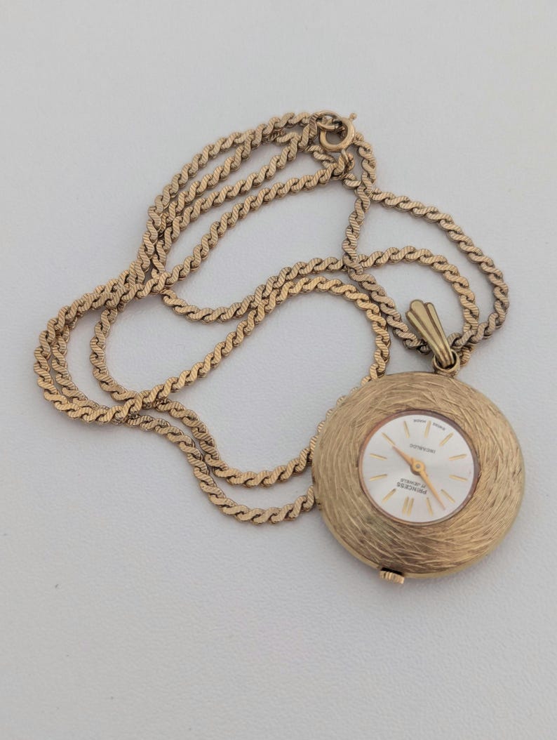 May include: A gold-toned pendant watch with a textured, circular face and a matching chain. The watch face has a white background with gold hands and markers. The chain is a twisted rope design.