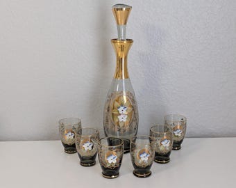 May include: A vintage glass decanter set with a tall, slender decanter and six matching glasses. The set features gold accents and floral designs with blue and white flowers. The decanter has a stopper, and the glasses have a dark base.