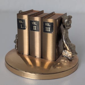 May include: A metallic gold-toned desk accessory featuring three upright book-shaped blocks labeled Vol. I, Vol. II, and Vol. III on black rectangular plaques. The set is mounted on a circular base and flanked by two small, sculpted metallic figures.