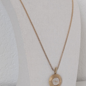 May include: Gold-toned pendant watch necklace. The watch face is white with black hands and markers. The pendant has a textured, circular design. The chain is a delicate, gold-colored rope style. The necklace is displayed on a white mannequin.