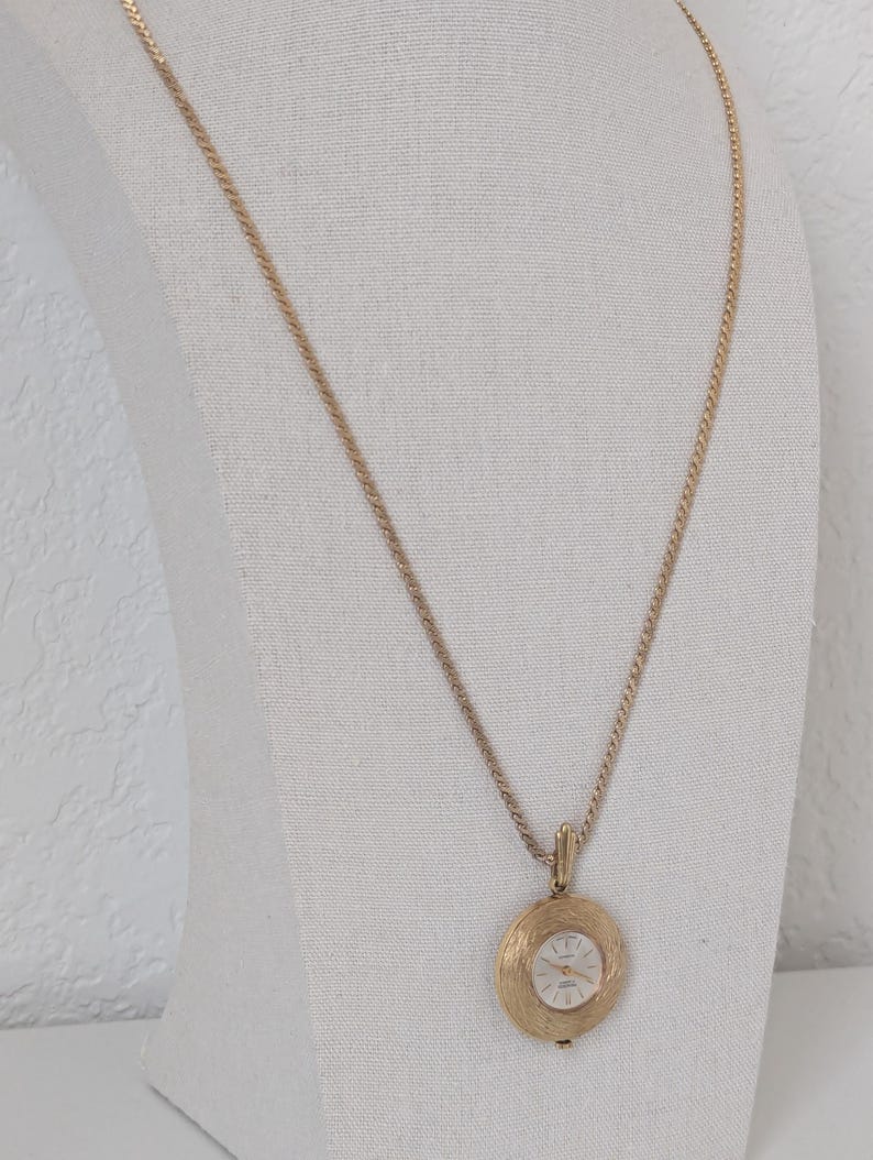 May include: Gold-toned pendant watch necklace. The watch face is white with black hands and markers. The pendant has a textured, circular design. The chain is a delicate, gold-colored rope style. The necklace is displayed on a white mannequin.