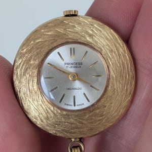 May include: A gold-toned Princess watch with a textured, circular face. The watch face is silver with black markers and hands. The text "PRINCESS 17 JEWELS INCABLOC SWISS MADE" is visible. A gold chain is attached to the watch.