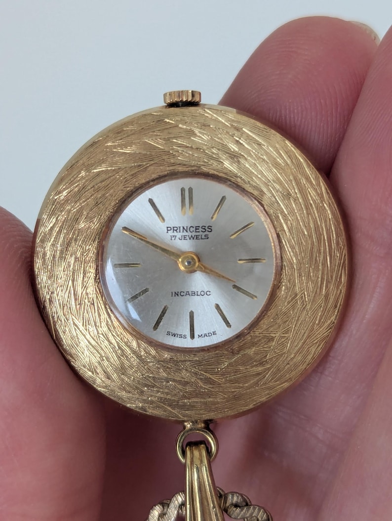 May include: A gold-toned Princess watch with a textured, circular face. The watch face is silver with black markers and hands. The text "PRINCESS 17 JEWELS INCABLOC SWISS MADE" is visible. A gold chain is attached to the watch.
