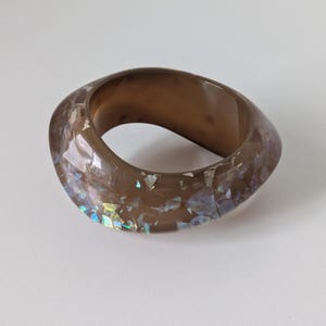 May include: A thick, translucent brown resin ring with an irregular, organic shape. The surface is embedded with small, reflective, iridescent flakes that shimmer with blue, green, and silver tones when catching the light, creating a textured appearance.