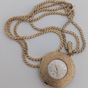 May include: A gold-toned pendant watch with a textured, circular face and a matching chain. The watch face is white with gold hands and markers, and the text "Princess Jewels" is visible. The chain is a twisted rope design.