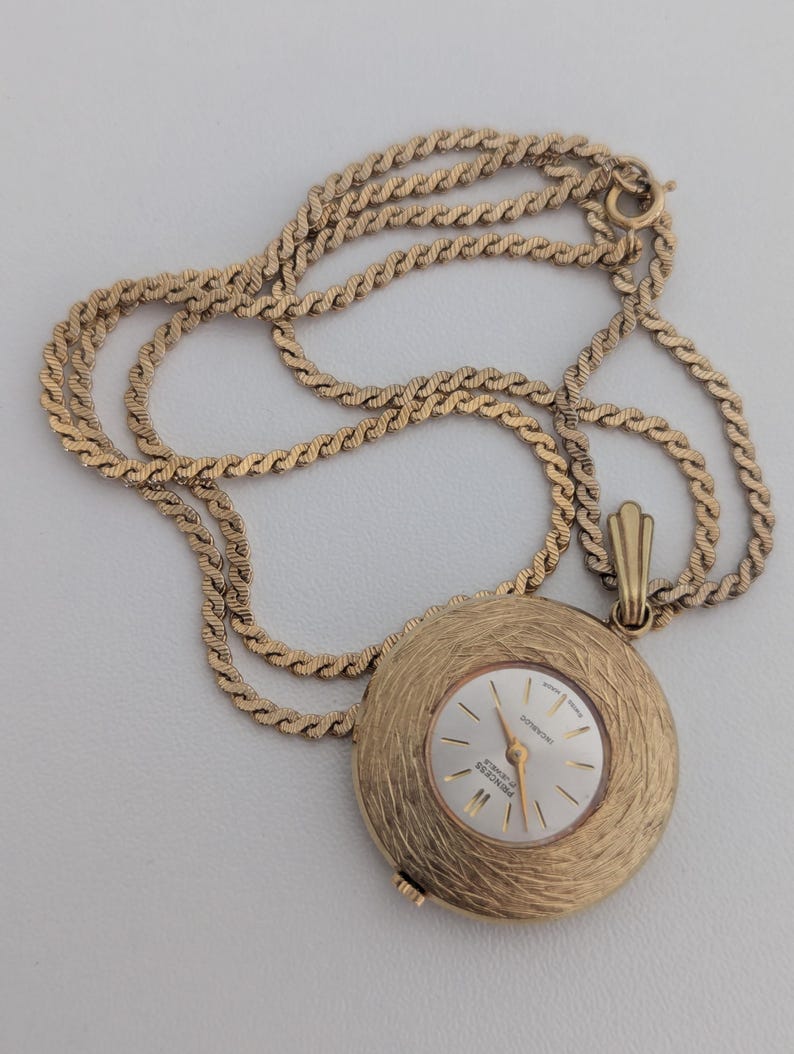 May include: A gold-toned pendant watch with a textured, circular face and a matching chain. The watch face is white with gold hands and markers, and the text "Princess Jewels" is visible. The chain is a twisted rope design.