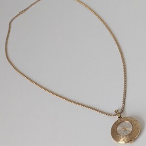 May include: A gold-toned necklace with a round pendant watch. The watch face has a light-colored background with gold-colored hands and hour markers. The watch is encased in a textured, circular frame. The chain is also gold-toned.