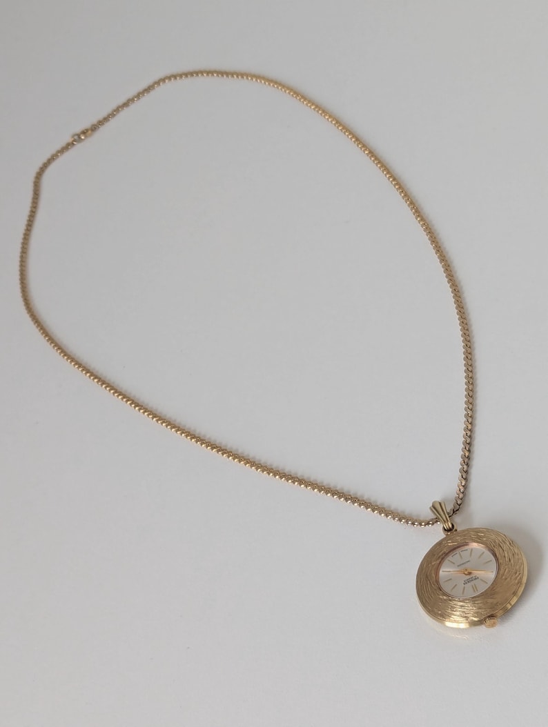 May include: A gold-toned necklace with a round pendant watch. The watch face has a light-colored background with gold-colored hands and hour markers. The watch is encased in a textured, circular frame. The chain is also gold-toned.