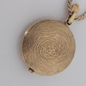 May include: A gold-colored, round pendant with a textured, spiral design. The pendant is attached to a gold-colored chain. The clasp is visible at the top, and the pendant appears to be closed. The background is a light gray color.