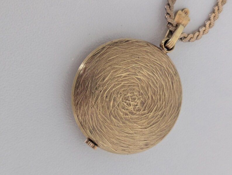 May include: A gold-colored, round pendant with a textured, spiral design. The pendant is attached to a gold-colored chain. The clasp is visible at the top, and the pendant appears to be closed. The background is a light gray color.