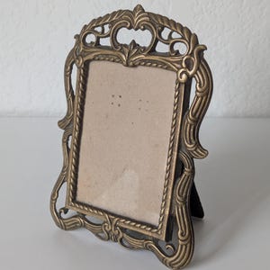 May include: A rectangular brass picture frame featuring an ornate, scrolled metal border with rope-like detailing. The frame has a vertical orientation and includes a stand on the back, holding a plain, light-colored insert within the central area.