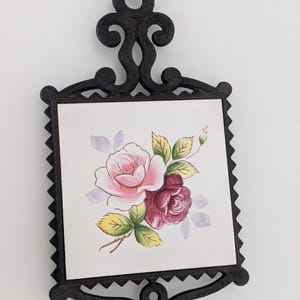 May include: A square trivet featuring a black cast iron frame with an ornate scrollwork handle and base. The center is a white ceramic tile decorated with a hand-painted floral motif showing a light pink rose and a deep red rose with green leaves.