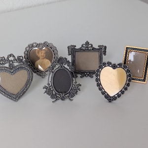 May include: A collection of six small decorative frames in various shapes including heart, oval, and rectangular designs. The frames feature ornate metallic finishes in silver, black, and gold tones, with intricate detailing along the edges.
