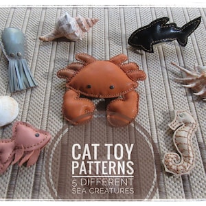 May include: Five leather cat toys shaped like sea creatures: a crab, a fish, an octopus, a seahorse, and a shark. The toys are arranged on a beige woven mat with the text "CAT TOY PATTERNS 5 DIFFERENT SEA CREATURES" in the centre.