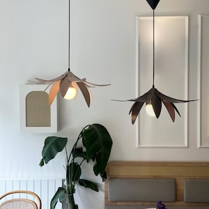 Peut inclure: Deux lampes suspendues en forme de fleur sont suspendues au plafond. Les lampes ont un sommet noir en forme de cône et un abat-jour en forme de fleur de couleur neutre et chaude. Elles surplombent un cadre de café avec une plante et une table.