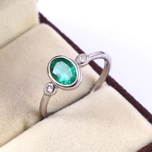 Puede incluir: Un anillo de plata con una esmeralda ovalada y dos diamantes redondos más pequeños en la banda.