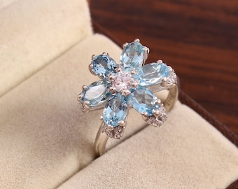 May include: A silver ring with a flower design featuring blue topaz gemstones and a smaller clear center stone.