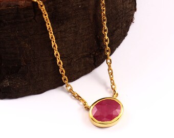 May include: A gold-colored necklace featuring a ruby-colored, oval-shaped gemstone pendant. The pendant is set in a gold bezel and hangs from a delicate gold chain. The necklace is displayed against a white background.