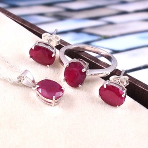 Op de afbeelding: Een sieradenset met een zilveren ring, een hangerketting en oorstekers. Elk stuk toont een levendige, ovaal geslepen robijn edelsteen. De ring en oorbellen hebben een eenvoudig ontwerp, terwijl de hanger aan een delicate ketting hangt.
