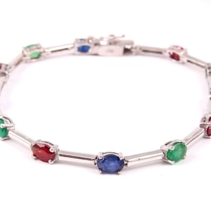 May include: A silver bracelet with alternating oval-shaped gemstones in red, green, and blue. The bracelet has a clasp closure.