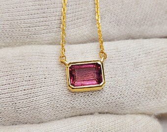 May include: A gold necklace featuring a rectangular, emerald-cut pink gemstone pendant set in a simple gold bezel. The pendant is suspended from a delicate gold chain, showcasing a vibrant, deep pink hue and a polished, reflective metal finish.